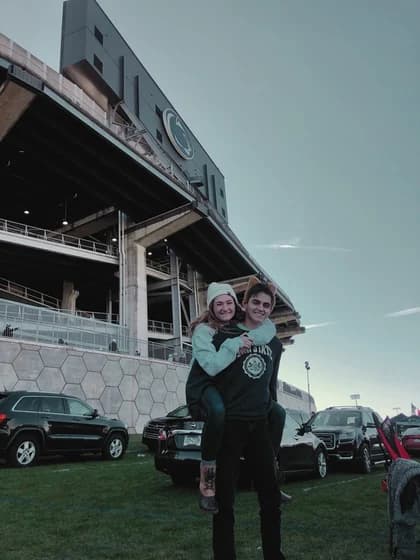 Allie and Alex outside a stadium at dusk, Allie on Alex's back, parked cars behind them.