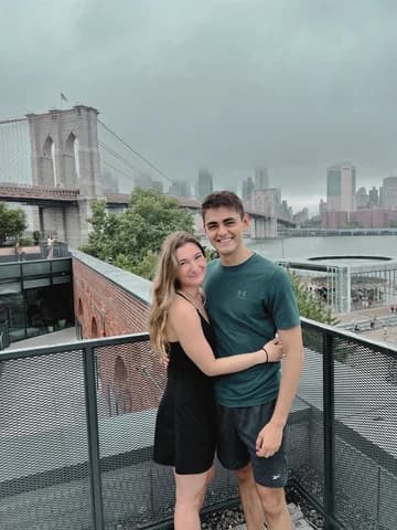 Allie and Alex on a railed walkway with the Brooklyn Bridge and the lower Manhattan skyline behind them.