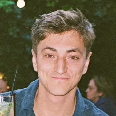 Best Man: a man with short light-brown hair in a dark blue polo shirt, holding a drink, warm string lights behind him.
