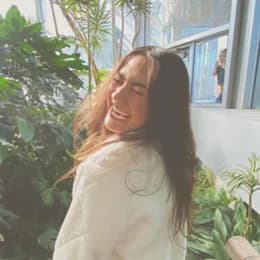 Bridesmaid: a woman with long dark hair in a cream knit sweater, laughing over her shoulder in a glasshouse.