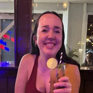 Bridesmaid: a woman with dark hair in a deep red top holding a tall cocktail, a restaurant lit blue and purple behind her.