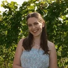 Maid of Honor: a woman with long dark hair in a pale blue floral dress, smiling, rows of grapevines behind her.